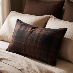 May include: A rectangular throw pillow featuring a dark brown and black plaid pattern. The pillow is positioned in front of two solid light beige pillows and behind a solid dark brown pillow, all resting on a light beige bed sheet.
