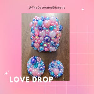 Può includere: Un set di tre oggetti decorativi, tra cui un quadrato e due pezzi rotondi, tutti ricoperti di perline rosa, blu e iridescenti. I pezzi presentano accenti a forma di cuore e sono etichettati "LOVE DROP".