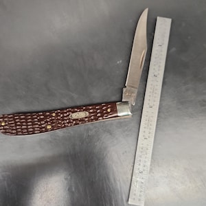 May include: A folding pocket knife with a dark brown, textured handle and a single silver-colored blade. The knife is positioned next to a metal ruler, showing the blade and handle length, with the word CASE visible on the handle and blade.