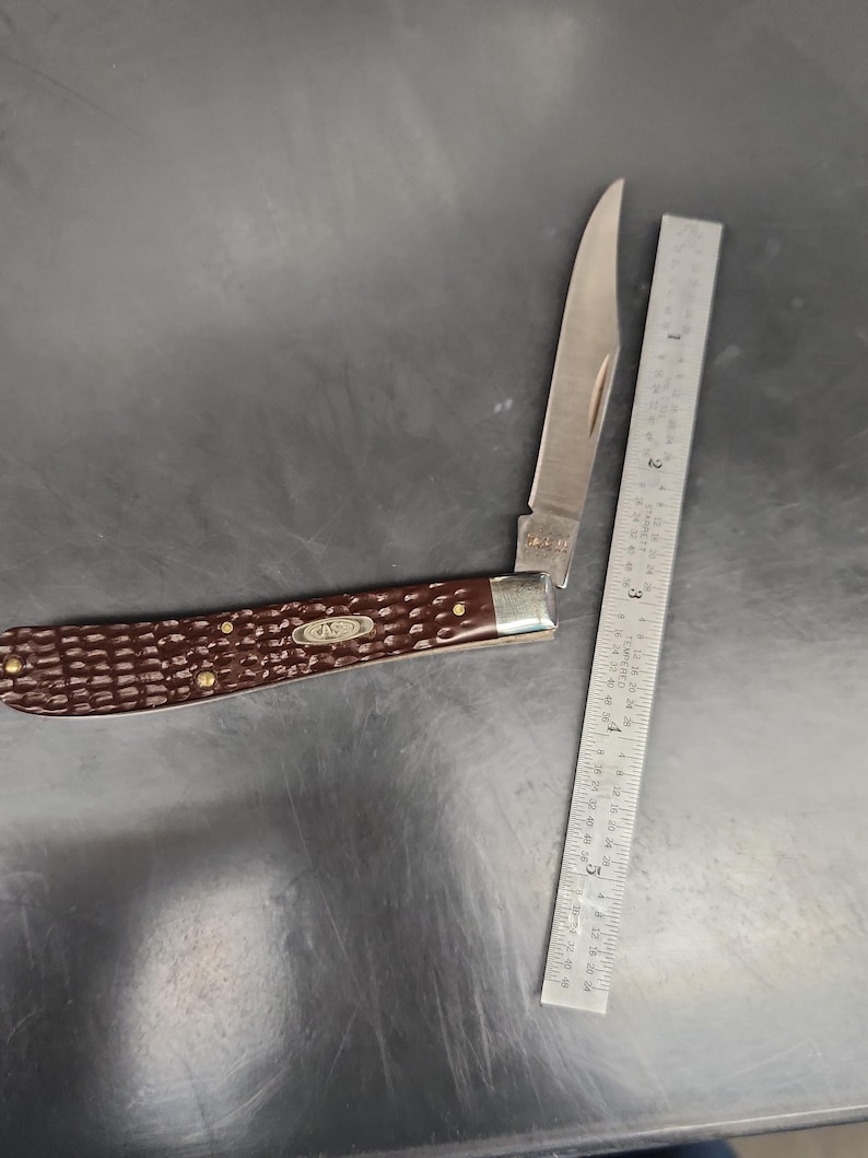 May include: A folding pocket knife with a dark brown, textured handle and a single silver-colored blade. The knife is positioned next to a metal ruler, showing the blade and handle length, with the word CASE visible on the handle and blade.