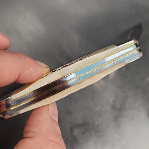 May include: A folding pocket knife with a cream-colored handle and metallic spacers. The handle features a textured surface with visible brass and steel layers along the spine. The knife is shown in a closed position with a metallic finish on the bolsters.
