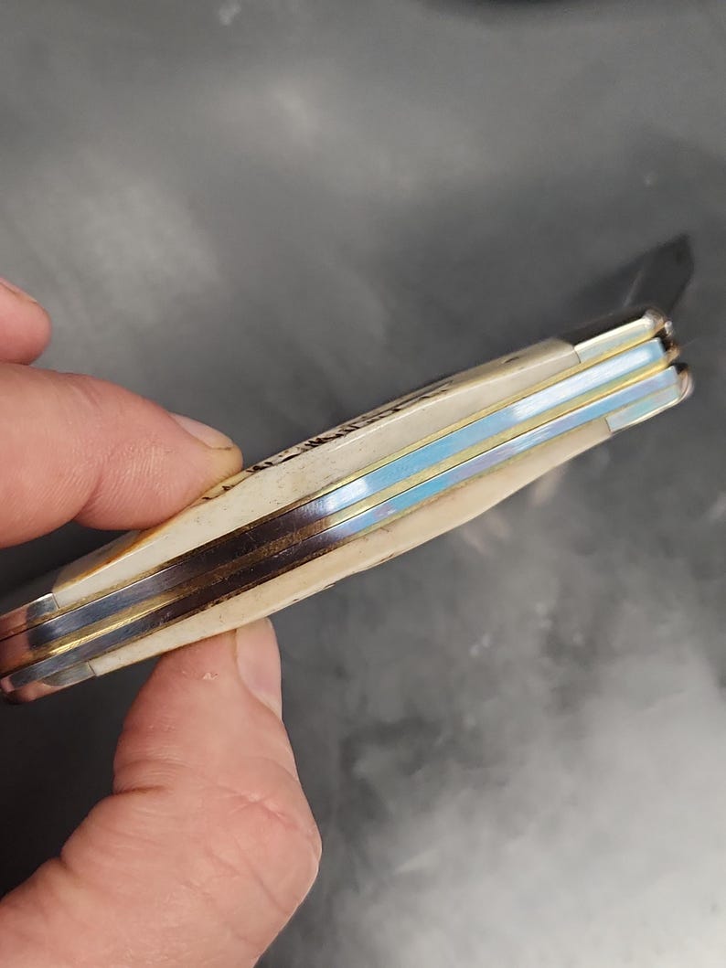May include: A folding pocket knife with a cream-colored handle and metallic spacers. The handle features a textured surface with visible brass and steel layers along the spine. The knife is shown in a closed position with a metallic finish on the bolsters.