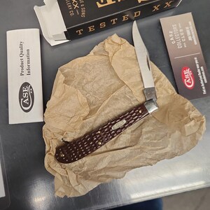 May include: A folding pocket knife with a textured dark brown handle and a silver blade, resting on crumpled tan paper. Included are a black and white Case XX box, a white product quality information card, and a Case Collectors Club card.