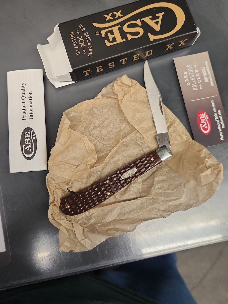 May include: A folding pocket knife with a textured dark brown handle and a silver blade, resting on crumpled tan paper. Included are a black and white Case XX box, a white product quality information card, and a Case Collectors Club card.