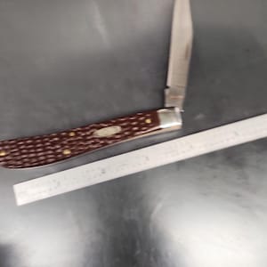 May include: A folding pocket knife with a dark brown, textured handle and a single silver-colored metal blade extended upward. The knife is positioned next to a metal ruler showing a length of approximately 4 inches for the handle section.