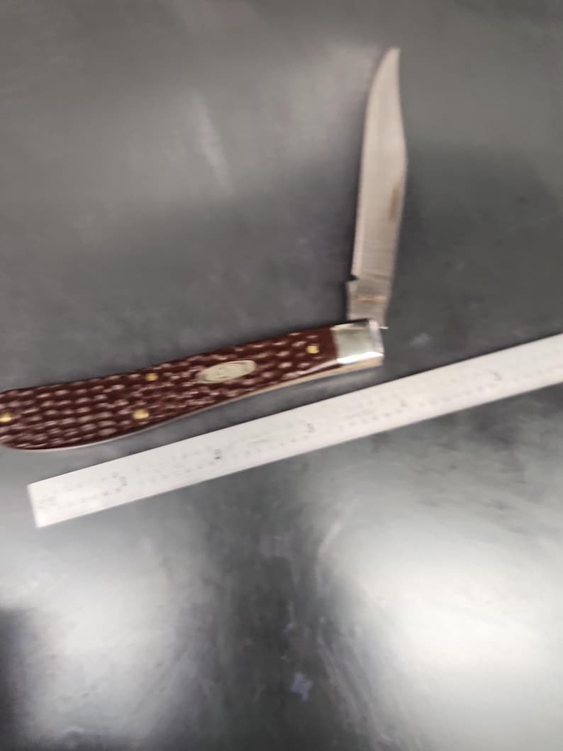 May include: A folding pocket knife with a dark brown, textured handle and a single silver-colored metal blade extended upward. The knife is positioned next to a metal ruler showing a length of approximately 4 inches for the handle section.
