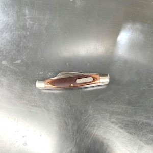 May include: A closed pocket knife with a brown handle and silver-colored metal bolsters at both ends. The handle features a silver-colored metal inlay with the text Old Timer engraved on it. The knife is in a folded position with the blade tucked inside.
