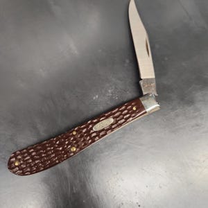 May include: A folding pocket knife with a single silver-toned blade and a dark brown handle featuring a textured, jigged pattern. The handle includes a silver metal bolster and a small oval shield with the embossed text CASE.
