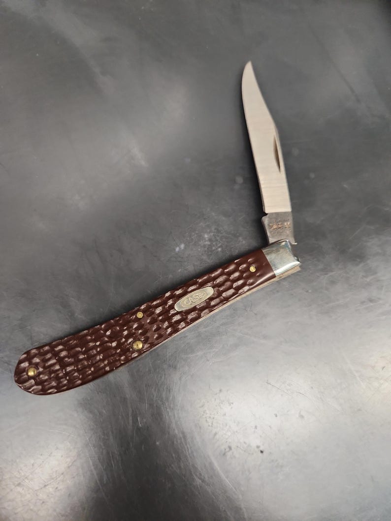 May include: A folding pocket knife with a single silver-toned blade and a dark brown handle featuring a textured, jigged pattern. The handle includes a silver metal bolster and a small oval shield with the embossed text CASE.
