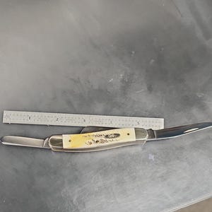 May include: A folding pocket knife with two silver-colored metal blades and a cream-colored textured handle. A metal ruler is positioned above the knife, showing the handle length is approximately 3.5 inches and the total length is 6 inches.