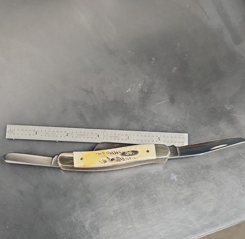 May include: A folding pocket knife with two silver-colored metal blades and a cream-colored textured handle. A metal ruler is positioned above the knife, showing the handle length is approximately 3.5 inches and the total length is 6 inches.
