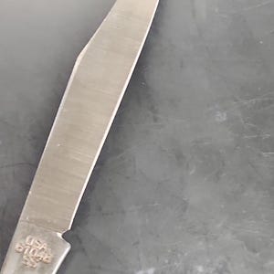 May include: A silver-colored metal knife blade with a pointed tip and a sharp edge. The base of the blade features an engraving that reads USA 61048 SS. The metal surface shows a brushed texture and reflects light along its length.