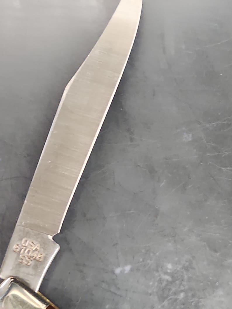 May include: A silver-colored metal knife blade with a pointed tip and a sharp edge. The base of the blade features an engraving that reads USA 61048 SS. The metal surface shows a brushed texture and reflects light along its length.