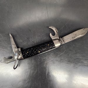 May include: A multi-tool pocket knife featuring a textured black handle with brass pins, a main blade, a bottle opener, a secondary blade, and a small metal loop attached to one end. The metal components show signs of wear and oxidation.