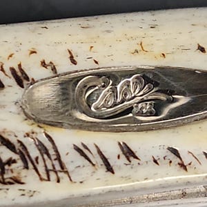 May include: A cream-colored handle with dark brown, irregular, linear markings. An oval-shaped silver-toned metal inlay is embedded in the center, featuring the embossed script text Case XX with a decorative flourish underneath the lettering.