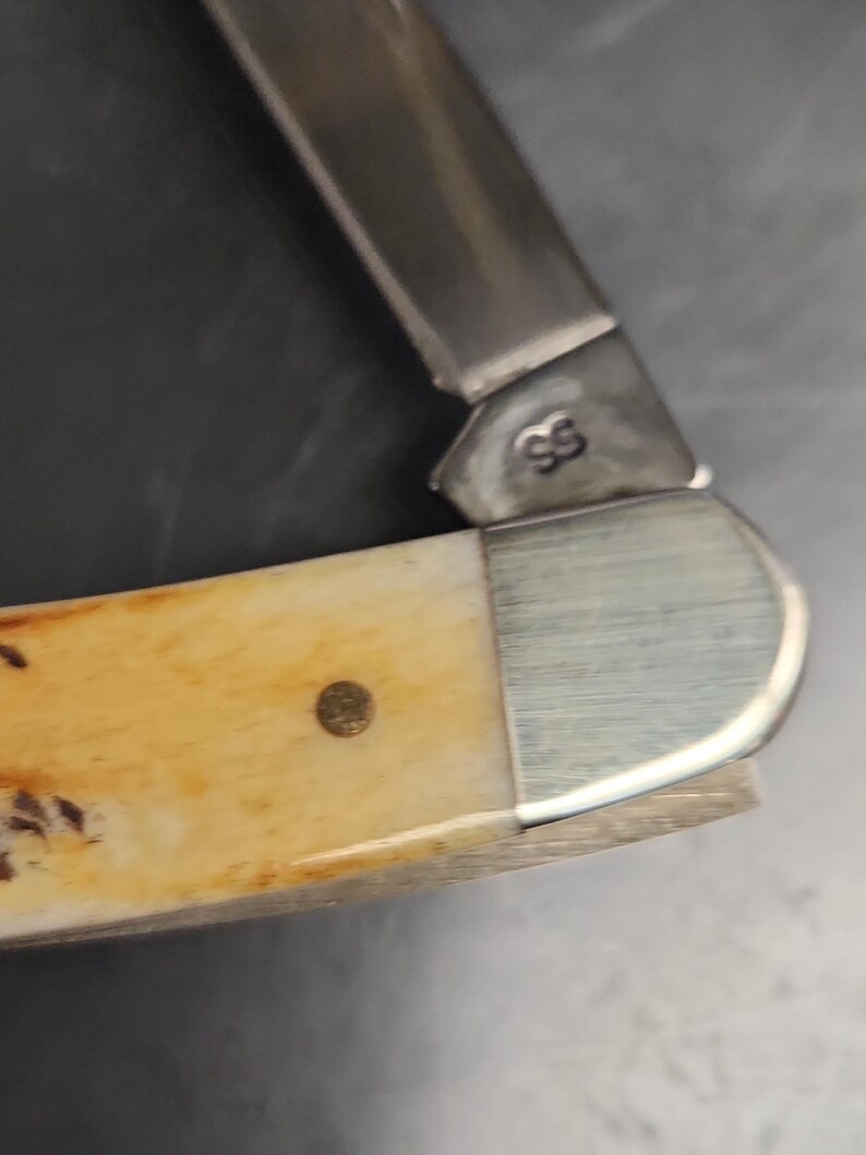 May include: A folding pocket knife featuring a cream-colored handle with brown mottled patterns and a single brass pin. The metal bolster is brushed silver, and the blade base is stamped with the letters SS in a clear, dark font.