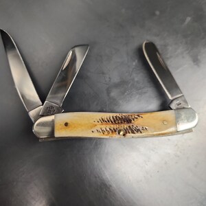 May include: A folding pocket knife with three stainless steel blades extended. The handle features a cream-colored bone texture with dark brown, feather-like markings and silver-toned metal bolsters at both ends. The blade base is stamped USA.