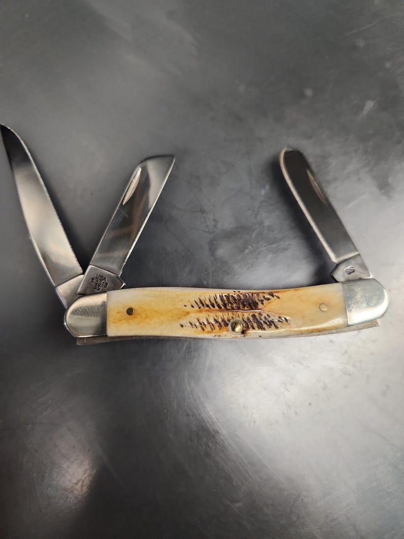 May include: A folding pocket knife with three stainless steel blades extended. The handle features a cream-colored bone texture with dark brown, feather-like markings and silver-toned metal bolsters at both ends. The blade base is stamped USA.