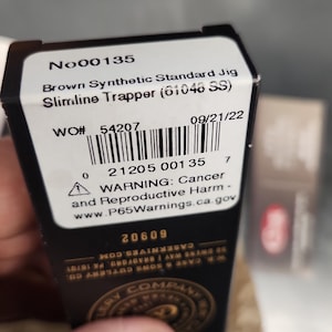 May include: A black rectangular product box with a white label featuring the text No00135, Brown Synthetic Standard Jig Slimline Trapper (61046 SS), a barcode, and a warning regarding cancer and reproductive harm with the website P65Warnings.ca.gov.