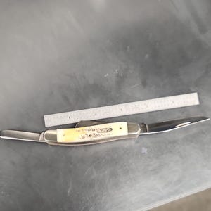 May include: A folding pocket knife with a cream-colored handle and two extended metal blades, positioned next to a metal ruler that shows the knife handle is approximately 3.5 inches long and the total length is about 6 inches.
