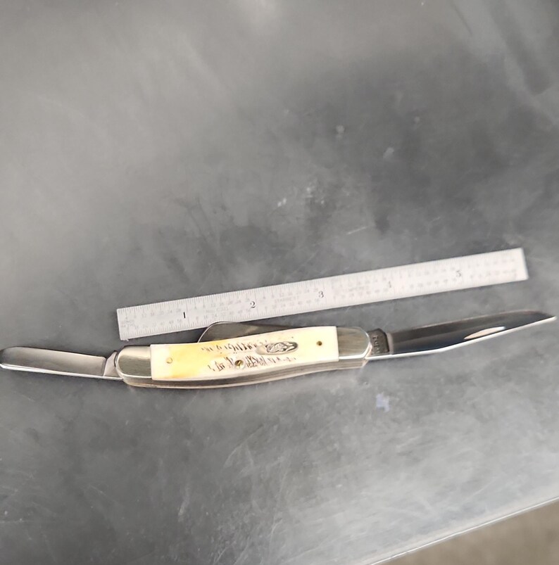May include: A folding pocket knife with a cream-colored handle and two extended metal blades, positioned next to a metal ruler that shows the knife handle is approximately 3.5 inches long and the total length is about 6 inches.