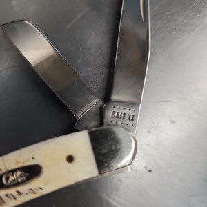 May include: A folding pocket knife with two open stainless steel blades and an off-white handle. The base of one blade is stamped with the text CASE XX, and the handle features a small metal inlay with the word Case embossed on the surface.