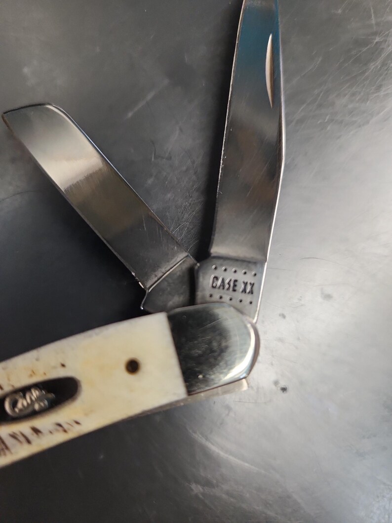 May include: A folding pocket knife with two open stainless steel blades and an off-white handle. The base of one blade is stamped with the text CASE XX, and the handle features a small metal inlay with the word Case embossed on the surface.