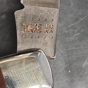 May include: A silver-colored metal knife blade featuring the stamped text CASE XX, positioned above a polished metal bolster. The blade surface displays a brushed texture, while the bolster reflects light with a smooth, metallic finish.