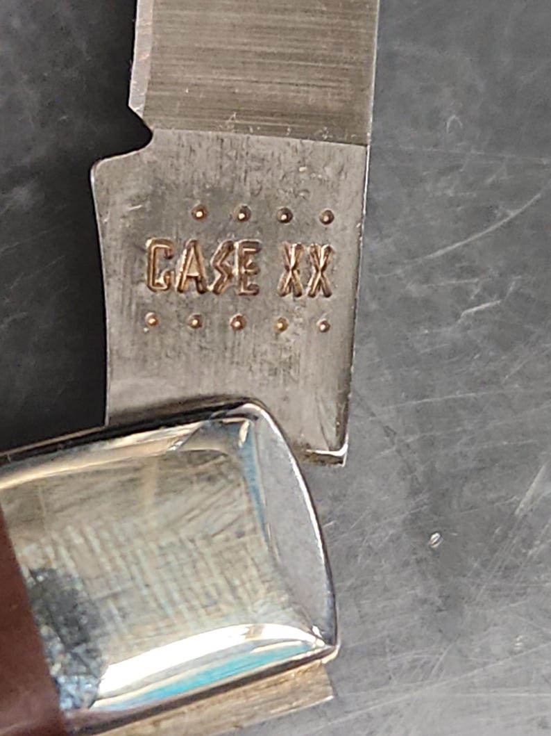 May include: A silver-colored metal knife blade featuring the stamped text CASE XX, positioned above a polished metal bolster. The blade surface displays a brushed texture, while the bolster reflects light with a smooth, metallic finish.