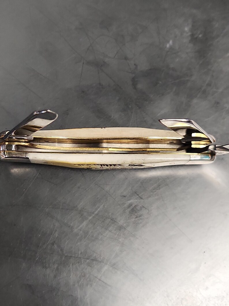 May include: A folding pocket knife with an off-white handle and metallic silver bolsters. The knife features multiple partially extended steel blades and internal brass liners visible between the handle scales, showing a multi-tool construction.