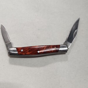 May include: A pocket knife with two blades extended. The handle is a reddish-brown wood grain, and the blades are silver. The knife is open, displaying both blades. The knife is on a white surface.