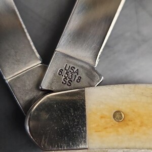 May include: A folding pocket knife with two metallic blades and a cream-colored handle. The base of one blade is stamped with the text USA 6.5318 SS. The knife features a polished metal bolster and a single brass rivet visible on the handle surface.