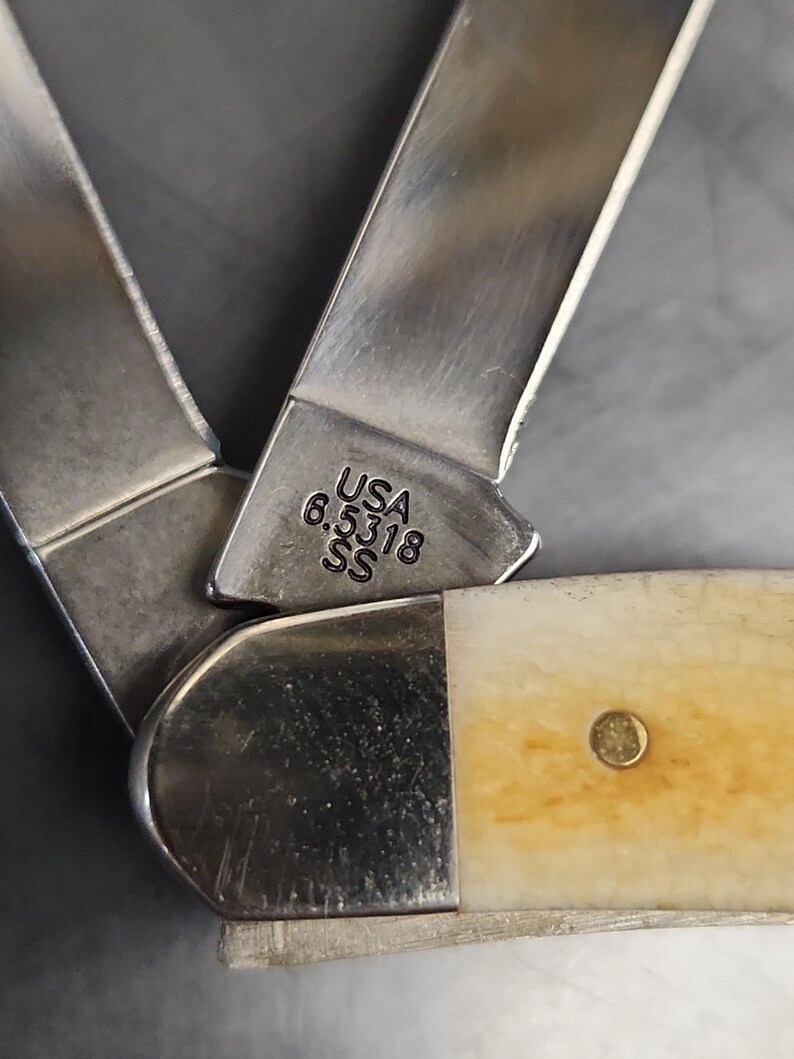May include: A folding pocket knife with two metallic blades and a cream-colored handle. The base of one blade is stamped with the text USA 6.5318 SS. The knife features a polished metal bolster and a single brass rivet visible on the handle surface.