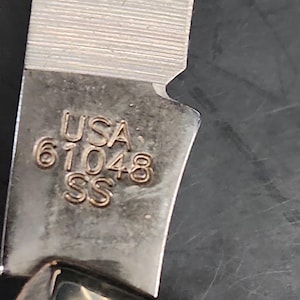 May include: A metallic knife blade featuring a stamped engraving that reads USA 61048 SS. The blade has a brushed finish with visible horizontal grain lines and a sharp edge, showing a clear transition from the tang to the sharpened cutting portion.