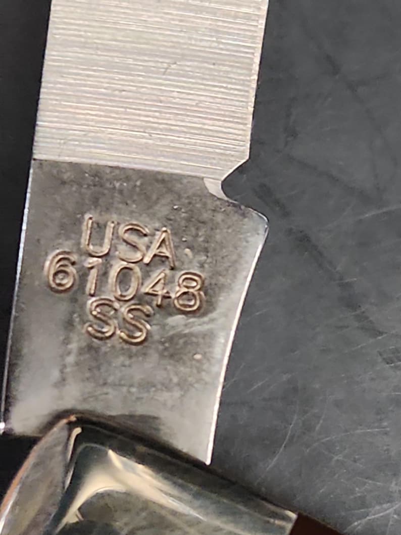 May include: A metallic knife blade featuring a stamped engraving that reads USA 61048 SS. The blade has a brushed finish with visible horizontal grain lines and a sharp edge, showing a clear transition from the tang to the sharpened cutting portion.