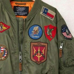 May include: Olive green bomber jacket with multiple embroidered patches. Patches include a Texas flag, a blue circle with a winged emblem, and a red shield with a dragon. The jacket has a zipper closure and brown leather zipper pulls.