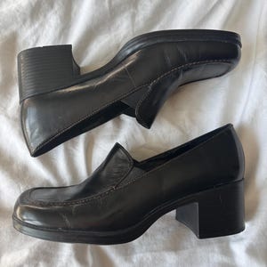 May include: A pair of black leather loafers featuring a square toe design, a thick block heel, and visible brown stitching along the vamp. The shoes have a slip-on construction with a smooth finish and a sturdy rubber sole for added traction.