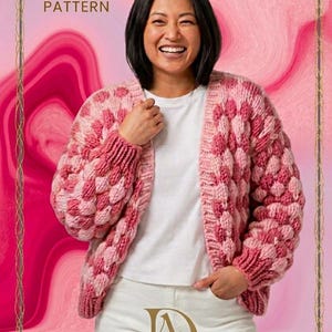 Könnte beinhalten: Eine Frau trägt einen rosa-weiß gestrickten Cardigan. Der Cardigan hat ein strukturiertes, blasenartiges Muster. Das Bild enthält den Text "COZY CABLE KNIT KNIT SWEATER PATTERN" und das Logo "DREAM & ADORE DESIGNS".