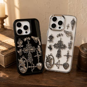 May include: Two smartphone cases, one black and one clear, each decorated with an assortment of silver-toned metal charms including ornate crosses, roses, a revolver, a bow and arrow, praying hands, and a heart-shaped pendant with a figure.