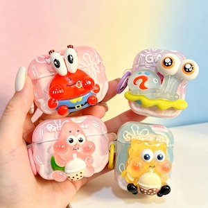 May include: Four protective cases for wireless earbuds featuring 3D character designs. The cases depict a red crab, a light blue snail, a pink starfish holding a drink, and a yellow sponge character holding a drink, all with glossy finishes.