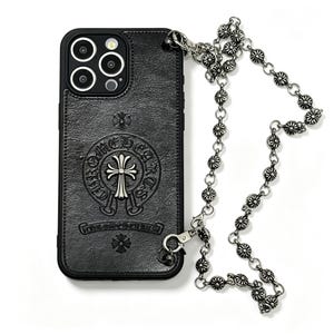 May include: A black textured phone case featuring a silver-toned cross emblem and the text Chrome Hearts embossed on the back. The case includes a long silver-toned metal chain strap with circular floral-patterned links attached via metal clips.