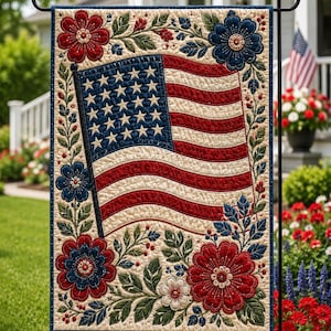 May include: A rectangular garden flag featuring a quilted design of the American flag centred within a cream-coloured border. The border is decorated with red and blue flowers, green leaves, and small red berries, all rendered in a stitched fabric style.