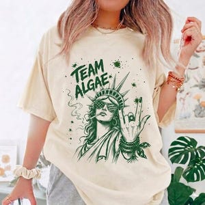 May include: A cream-colored short-sleeve t-shirt featuring a dark green graphic print of the Statue of Liberty wearing sunglasses, smoking, and making a rock-and-roll hand gesture. The text "TEAM ALGAE" is printed above the figure in green.