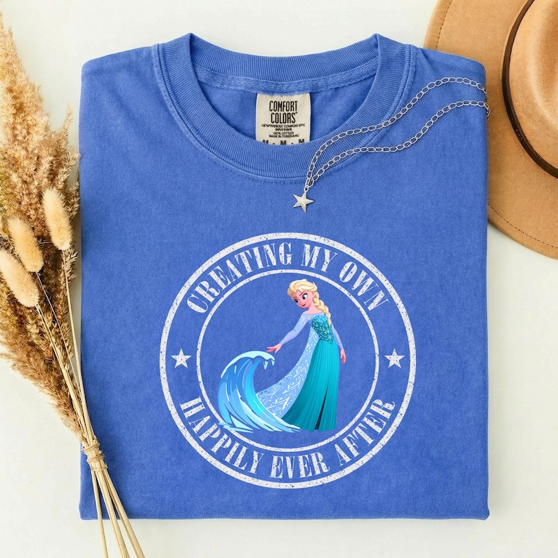 Puede incluir: Camiseta azul Comfort Colors con un gr&aacute;fico circular con un personaje de dibujos animados y el texto "CREATING MY OWN HAPPILY EVER AFTER". Un collar con colgante de estrella plateada descansa sobre la camisa.