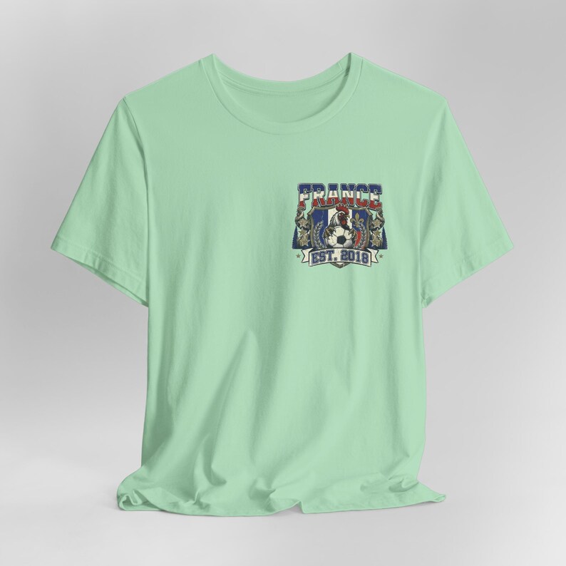 May include: A light mint green short-sleeve t-shirt featuring a graphic on the left chest. The design includes a rooster, a soccer ball, and the text "FRANCE" and "EST. 2018" in blue, white, and red colors, framed by decorative elements.