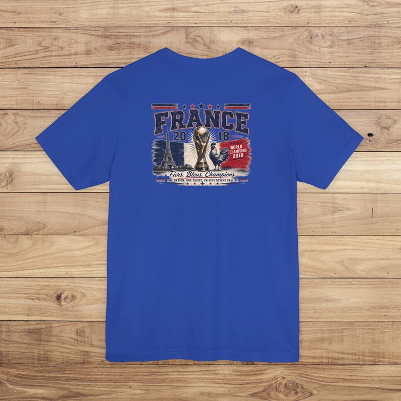 May include: A royal blue short-sleeve t-shirt featuring a graphic on the back. The design includes the text FRANCE 2018, an illustration of the Eiffel Tower, the World Cup trophy, a rooster, and the text Fiers. Bleus. Champions.