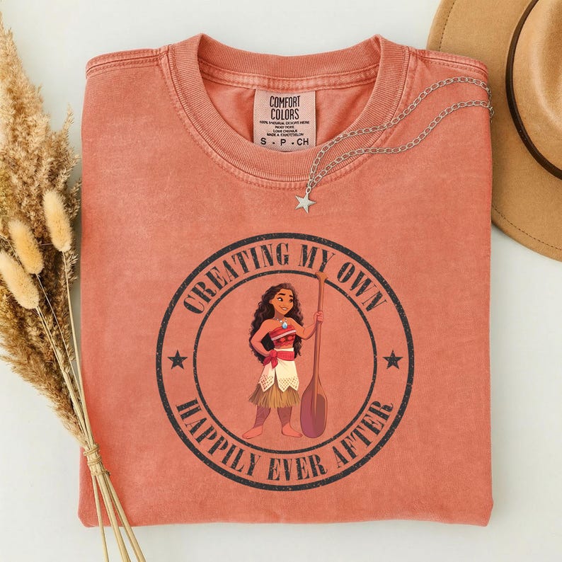 Op de afbeelding: Koraalrood T-shirt met een afbeelding van een vrouw met een peddel, omringd door de tekst "CREATING MY OWN HAPPILY EVER AFTER". Een zilveren sterrenketting en een beige hoed zijn ook zichtbaar. Het shirt heeft een Comfort Colors label.