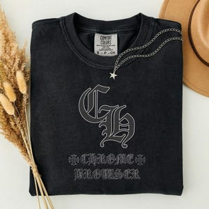 May include: A black crewneck sweatshirt featuring a large grey gothic-style monogram of the letters CH. Below the monogram, the text CHROME BROWSER is printed in a matching gothic font, flanked by two small cross symbols. A silver star necklace rests on top.