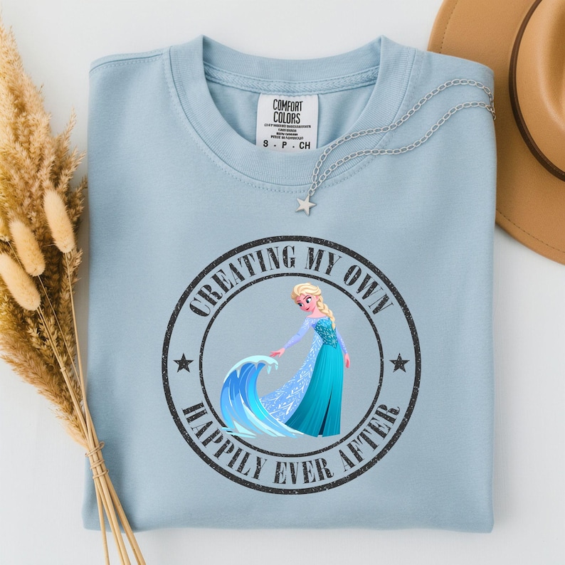 Puede incluir: Camiseta Comfort Colors azul claro con cuello redondo. La camiseta presenta un gr&aacute;fico de un personaje de dibujos animados con un vestido azul, con el texto "CREATING MY OWN HAPPILY EVER AFTER" en un dise&ntilde;o circular. Un collar plateado con un colgante de estrella est&aacute; en la camiseta.