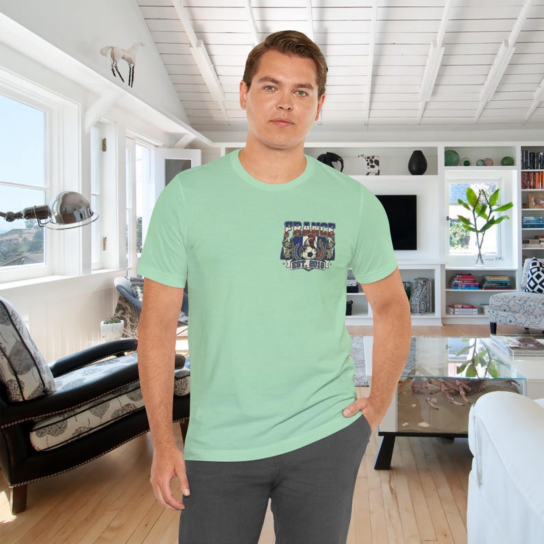 May include: A light green short-sleeve t-shirt featuring a small graphic on the left chest. The graphic displays a soccer ball, a rooster, and the text "FRANCE" and "EST. 2019" in blue, white, and red colors. The shirt has a crew neckline.